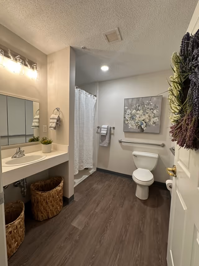 Well-lit assisted-living bathroom with sink and mirror, shower with white curtain, toilet with grab bar, floral wall art, and wicker baskets.