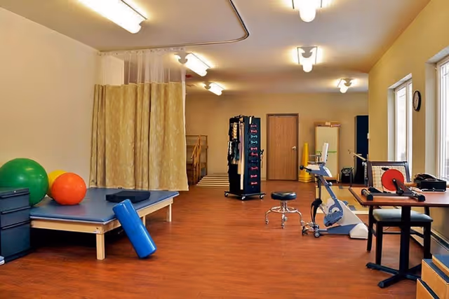 Spacious rehab/exercise therapy room with a treatment table, exercise balls, stationary bike, weights, and a privacy curtain.