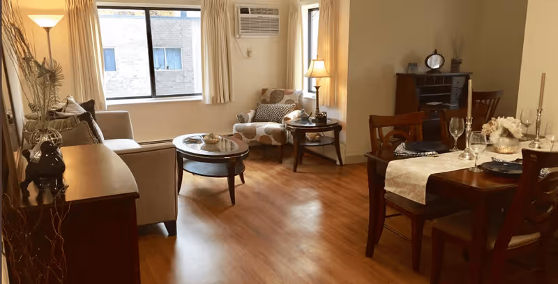 A cozy senior living room and dining area with wooden flooring. The living room features a beige sofa, a patterned armchair, a round coffee table, and a side table with a lamp. The dining area has a wooden table set with plates, wine glasses, and candles. A window with cream curtains and an air conditioning unit is visible in the background.