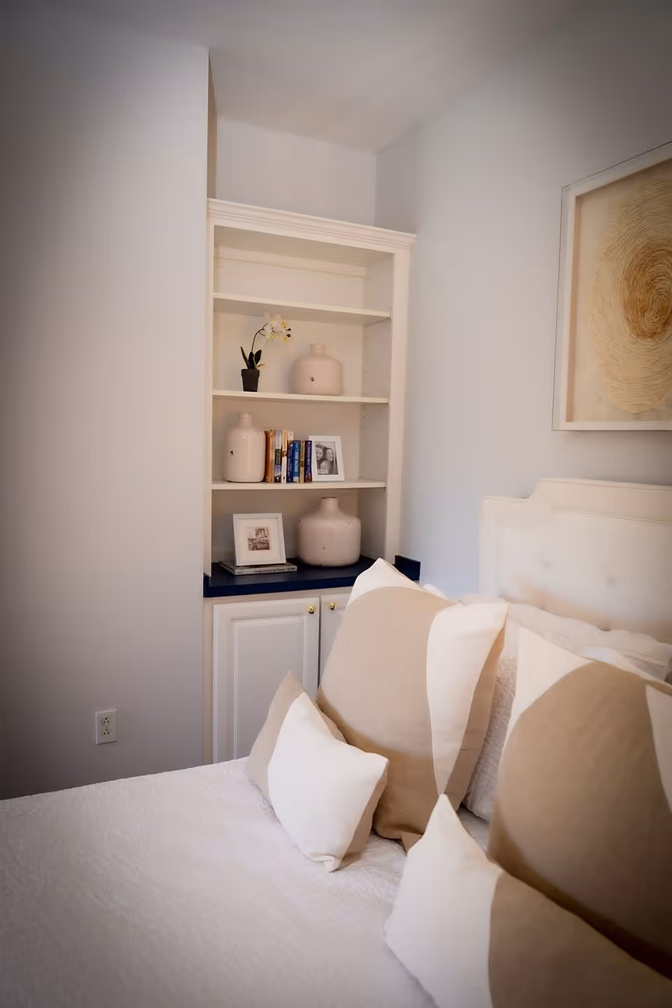 A cozy bedroom corner featuring a white bed with multiple beige and white pillows. Next to the bed is a built-in white shelving unit with decorative vases, books, and framed photos. A framed abstract artwork hangs on the wall above the bed.