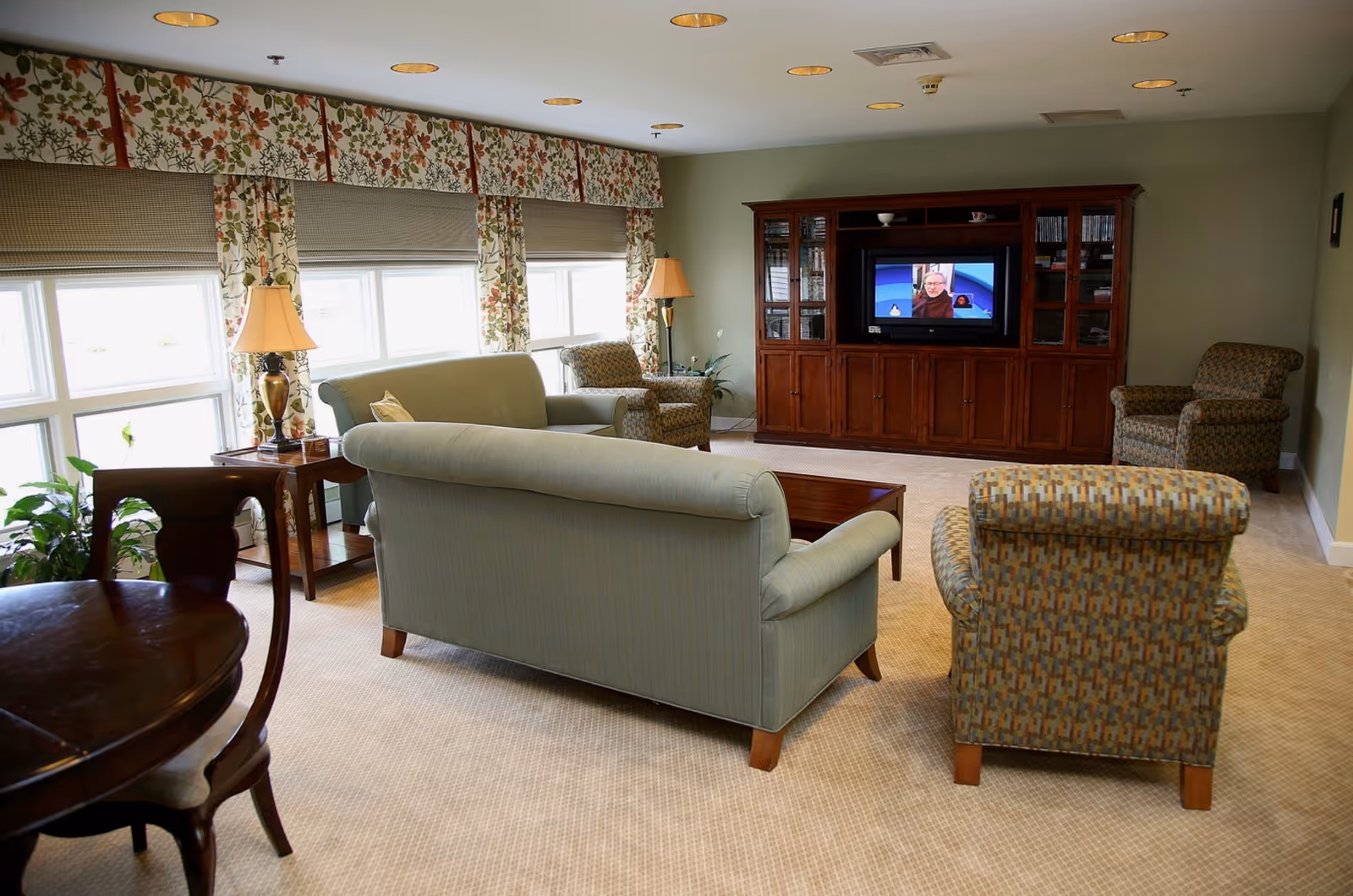 A cozy living room with light green sofas and patterned armchairs arranged around a wooden coffee table. There is a large wooden entertainment center with a television showing a news program. The room has large windows with floral curtains and beige carpeted flooring. A wooden dining table and chairs are partially visible in the foreground.