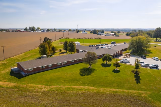 Aerial view of Falls City Nursing and Rehabilitation Center, a single-story building with a large parking lot surrounded by green lawns and trees, set in a rural area with fields and farm buildings in the background under a clear sky.