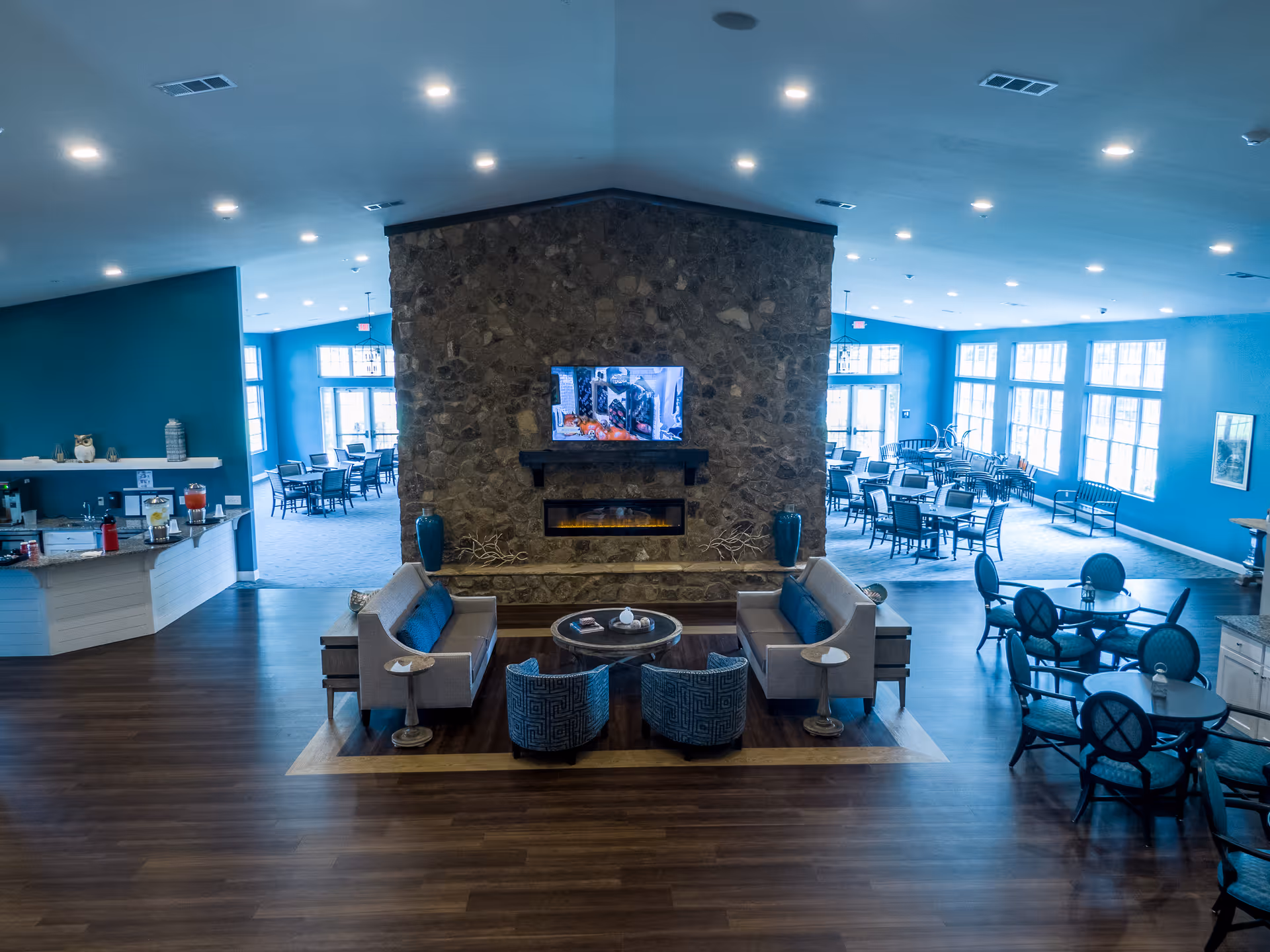 A spacious and well-lit common area in an assisted living facility featuring a stone fireplace with a mounted TV above it. The seating area includes two sofas, two armchairs, and small side tables arranged on a rug. Surrounding the seating area are multiple dining tables and chairs, with large windows allowing natural light to fill the room. The walls are painted blue, and there is a counter area with beverages on the left side.
