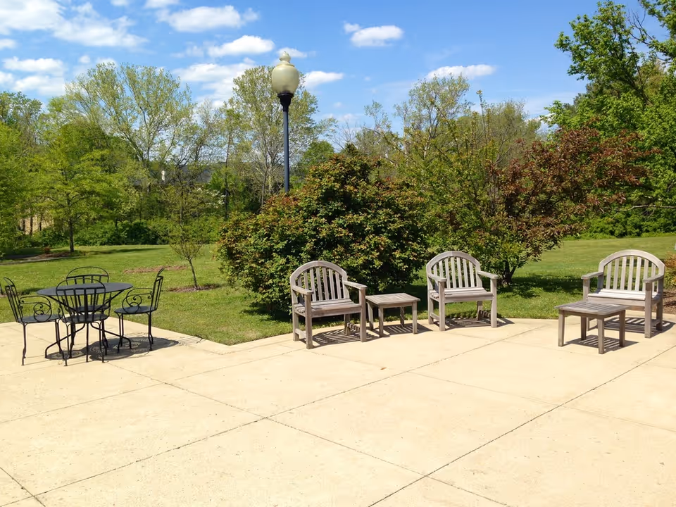 Outdoor patio area with metal table and chairs on the left and wooden chairs and small tables on the right, surrounded by green grass, bushes, and trees under a blue sky with scattered clouds.