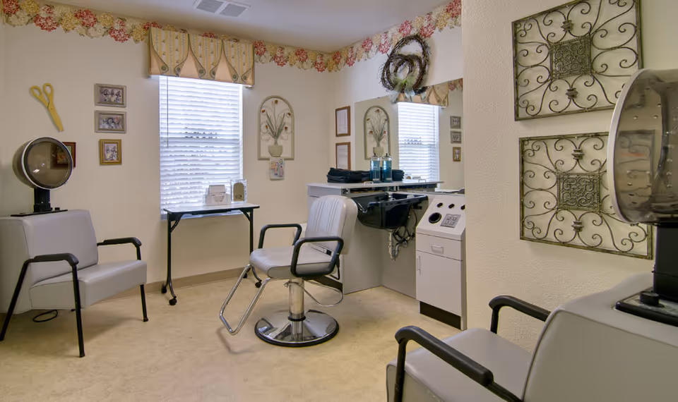 A small salon room with two salon chairs, a hair dryer, a styling station with a mirror, and decorative wall art. The room has two windows with blinds and floral valances, beige walls, and a light-colored floor.