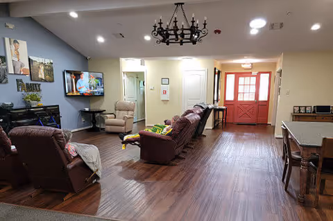 Interior view of a living room area in an assisted living facility with several brown recliner chairs arranged facing a wall-mounted TV. The room has wood flooring, a chandelier hanging from the ceiling, and a red door at the far end. There is a small table with chairs on the right side and family-themed wall decor on the left wall.