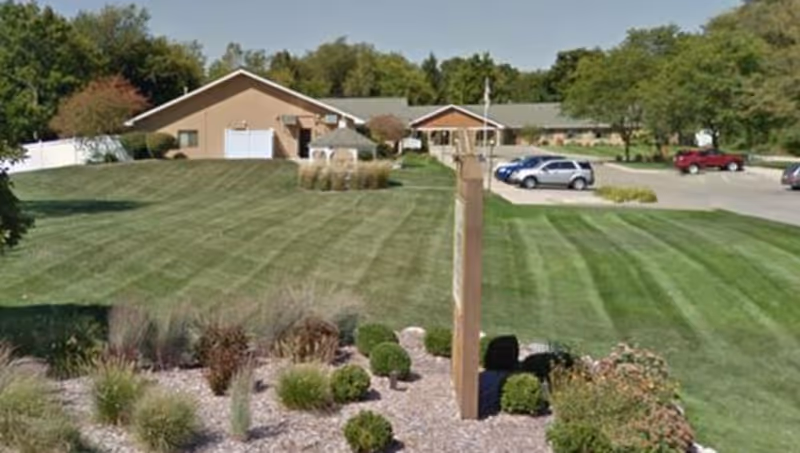 View of a senior living facility named Woodside with a large, well-maintained lawn and landscaped garden in the foreground. The building is a single-story structure with a beige exterior and a parking lot with several cars on the right side. Trees and greenery surround the area under a clear sky.