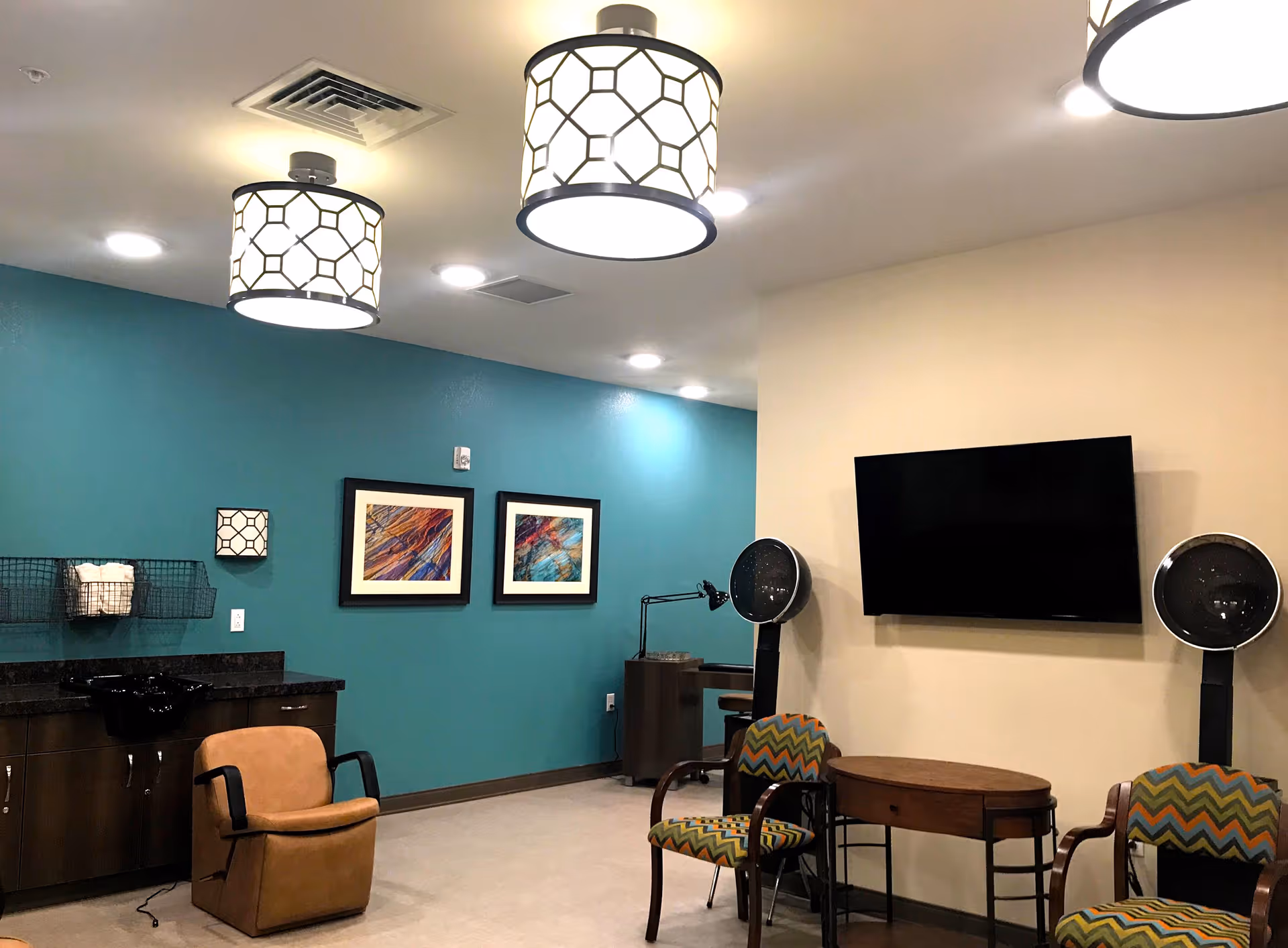 Interior common room with salon chairs and hair dryers, a wall-mounted TV, and colorful artwork on a teal accent wall.