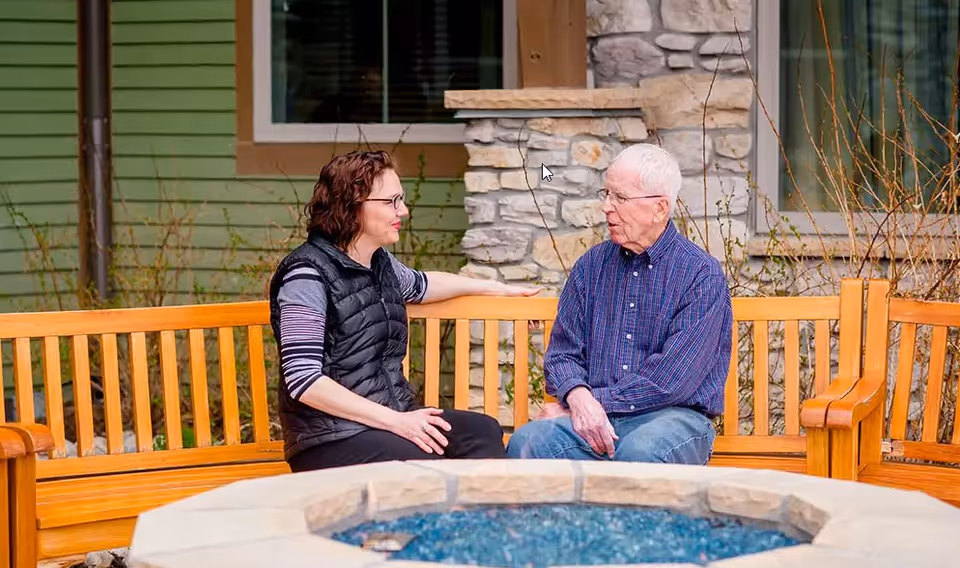 A woman and an elderly man sit on a wooden bench talking outside near a stone fire pit in front of a building.