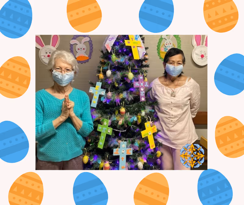 Two elderly women wearing face masks standing on either side of a decorated Christmas tree with colorful crosses and Easter egg ornaments. The background wall is decorated with bunny face crafts made from paper plates.