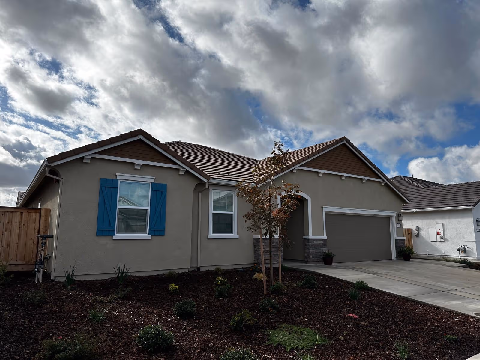 Front exterior of a single-story suburban house with a two-car garage, blue window shutters, and a landscaped front yard under a cloudy sky.