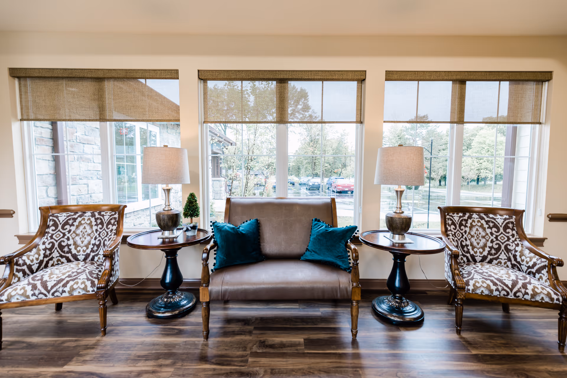 A sitting area with a leather loveseat flanked by patterned armchairs, side tables with lamps, and large windows behind.