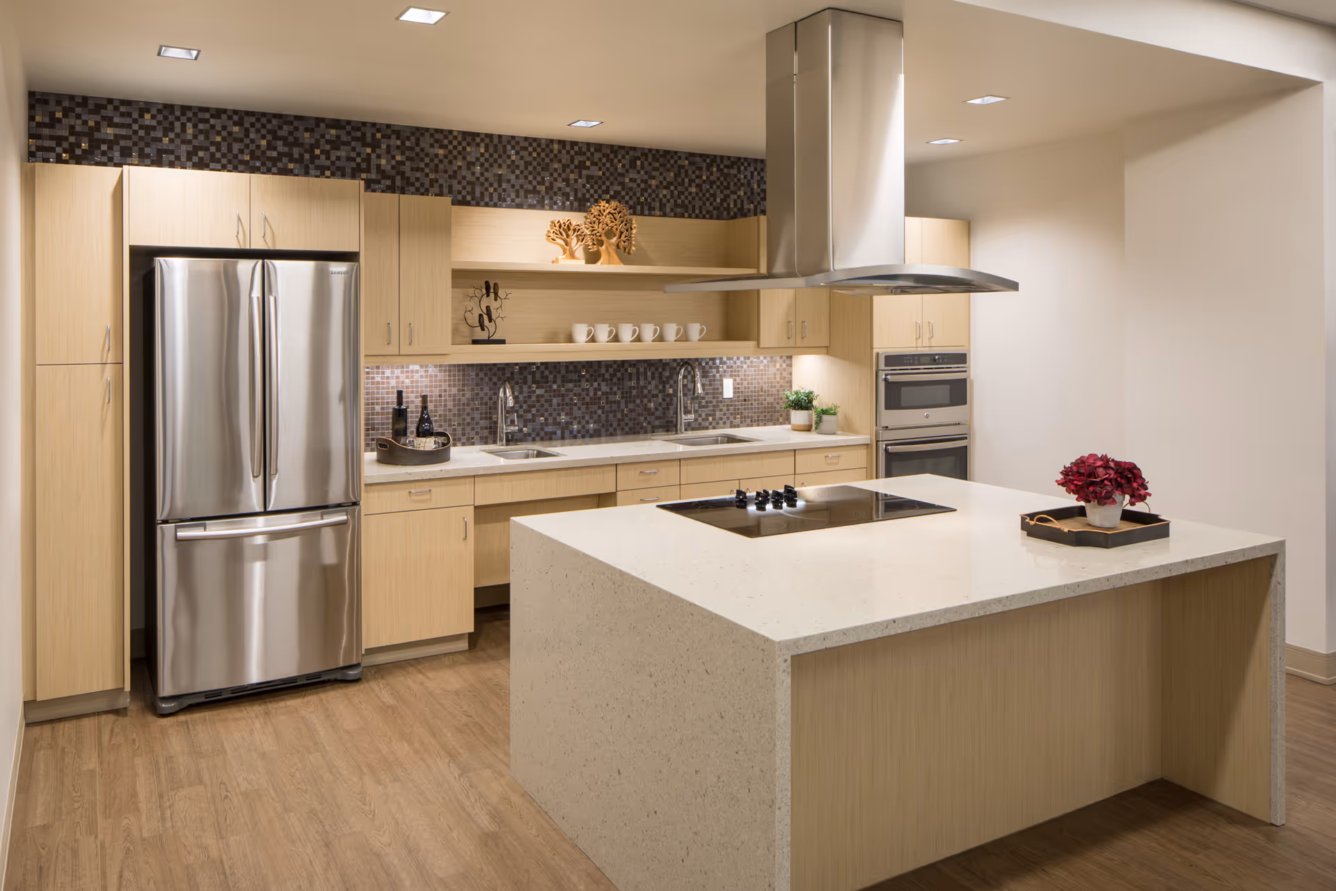 Modern kitchen with light wood cabinets, a large island with a built-in stovetop, stainless steel refrigerator, double sinks, and a mosaic tile backsplash. The kitchen is well-lit with recessed lighting and decorated with small plants, cups, and decorative items.