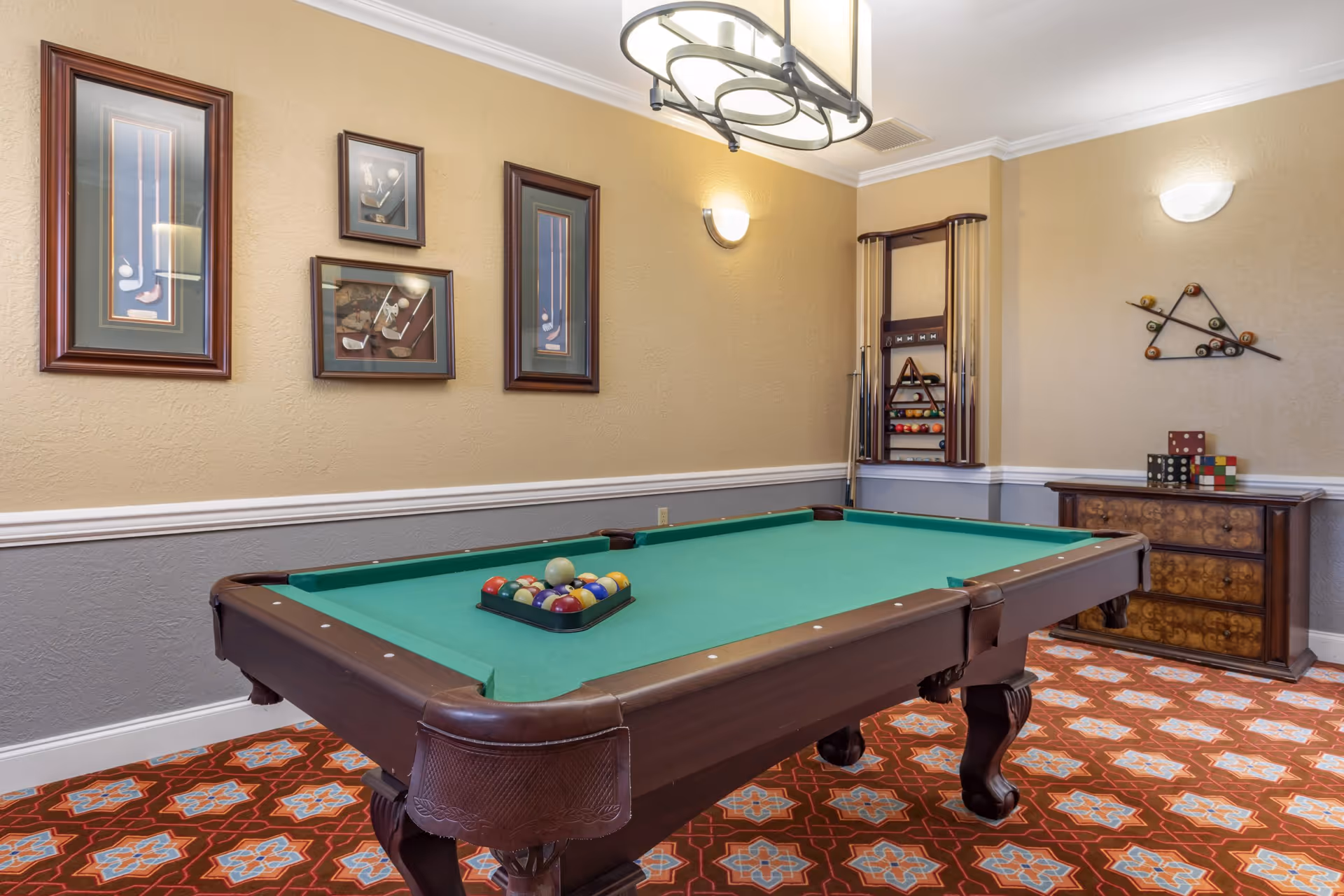 A game room with a green felt pool table set up for a game, with pool cues and racks on the wall. The room has beige and gray walls with framed pictures of golf equipment, a patterned carpet, and a wooden chest with decorative items on top.