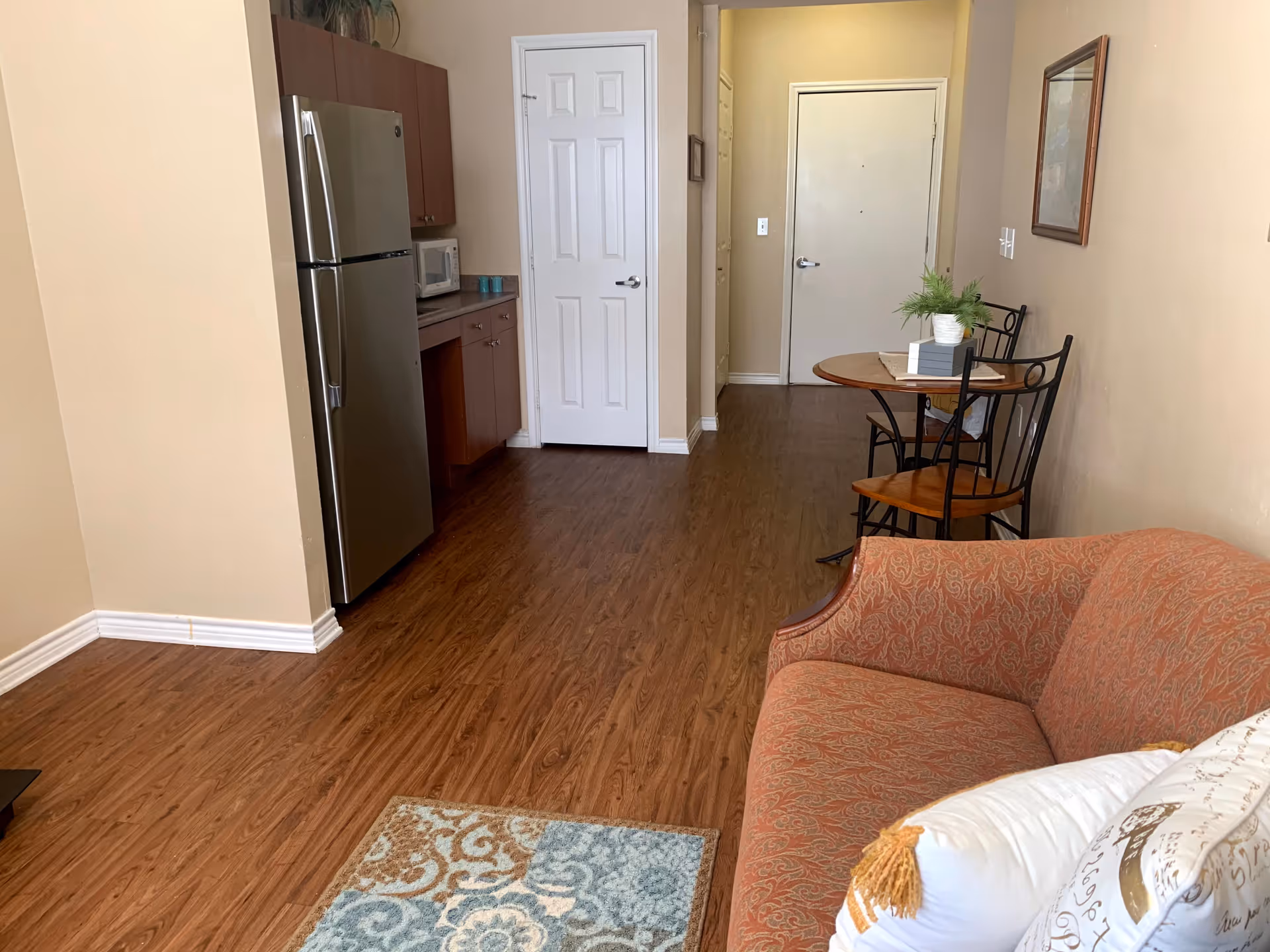 Interior view of a senior living facility apartment showing a small kitchen area with a stainless steel refrigerator, microwave, and wooden cabinets on the left. A hallway leads to a white door at the end. On the right side, there is a small round wooden table with two chairs and a potted plant on top. In the foreground, there is an orange patterned sofa with decorative pillows and a small area rug on the wooden floor.
