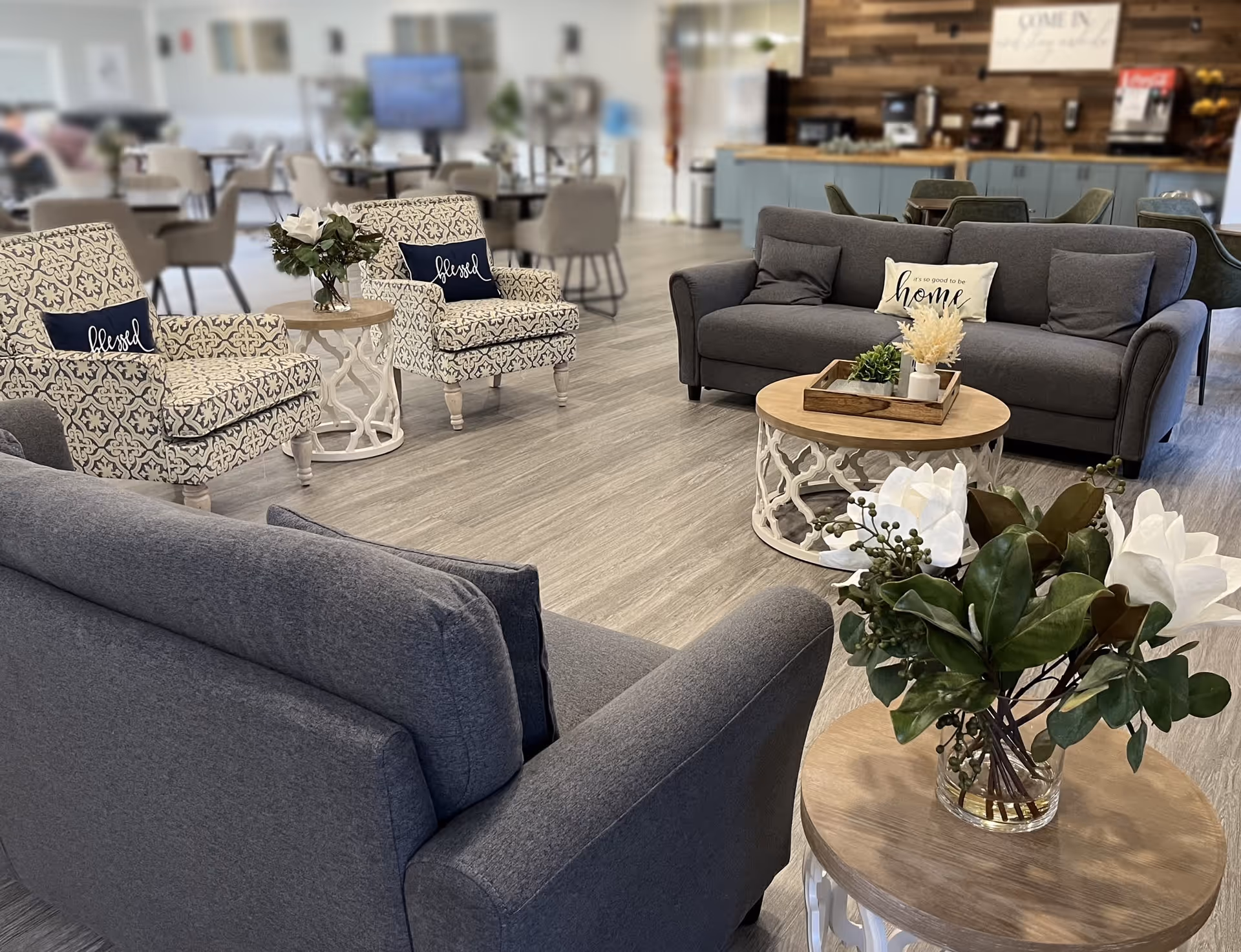 A cozy and well-decorated living room area in an assisted living community featuring two gray sofas and two patterned armchairs with 'blessed' pillows. There are round wooden coffee tables with decorative plants and flowers. In the background, there are dining tables and chairs, a TV, and a counter area with a coffee machine and a sign that says 'Come in, stay awhile.'