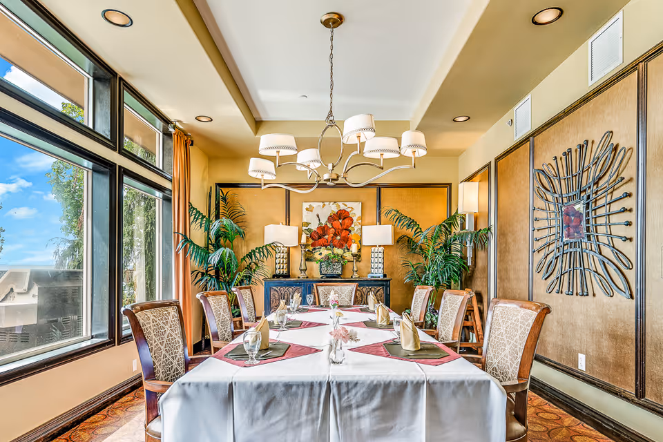 A well-lit dining room with a large rectangular table covered with a white tablecloth and set with glasses, napkins, and placemats. The room features large windows on the left side letting in natural light, decorative plants, a chandelier with multiple lampshades hanging above the table, and artwork on the walls including a floral painting and a metal wall sculpture.