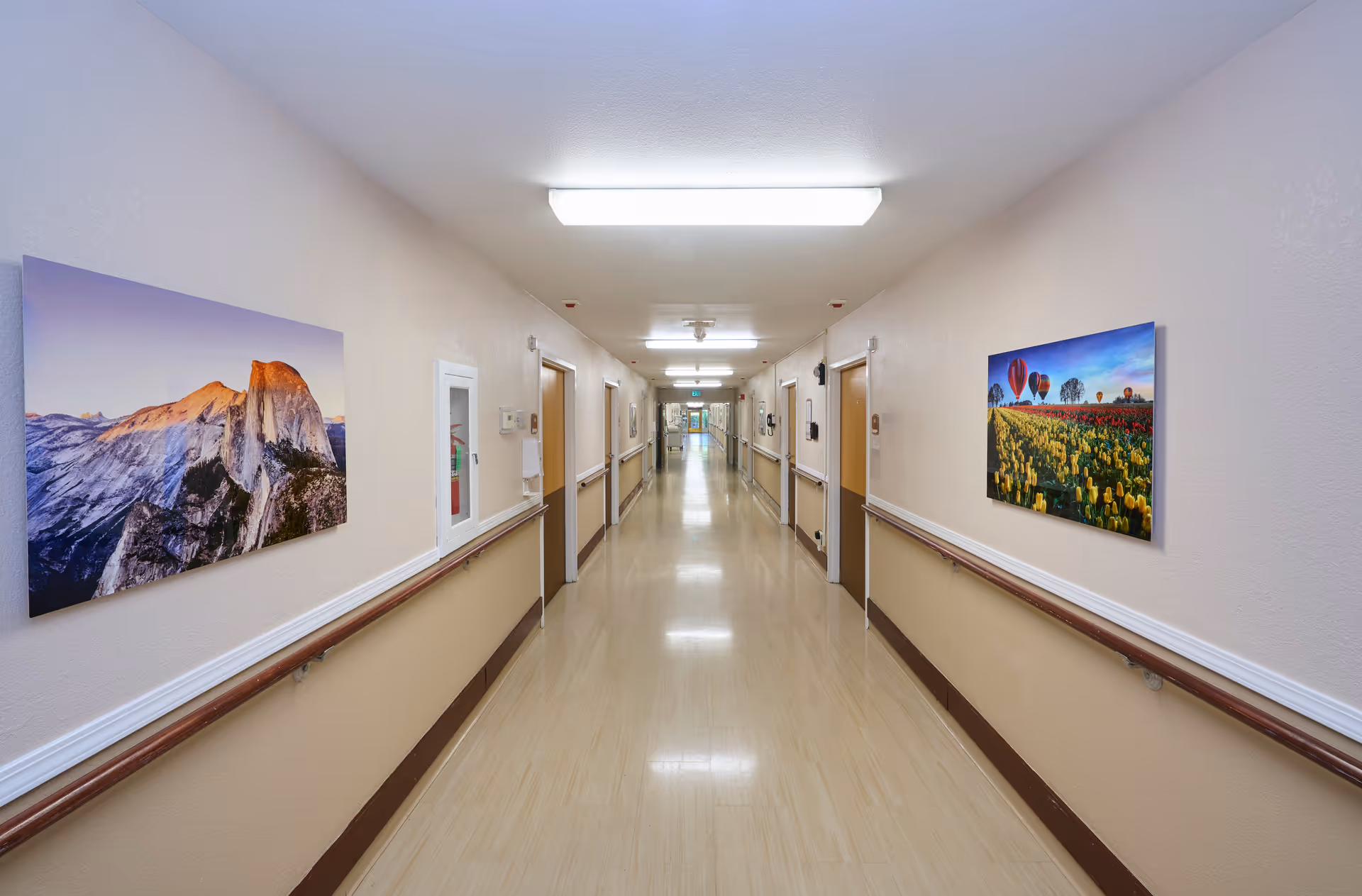 Long, well-lit interior hallway in a care facility with handrails and framed landscape pictures on the walls.