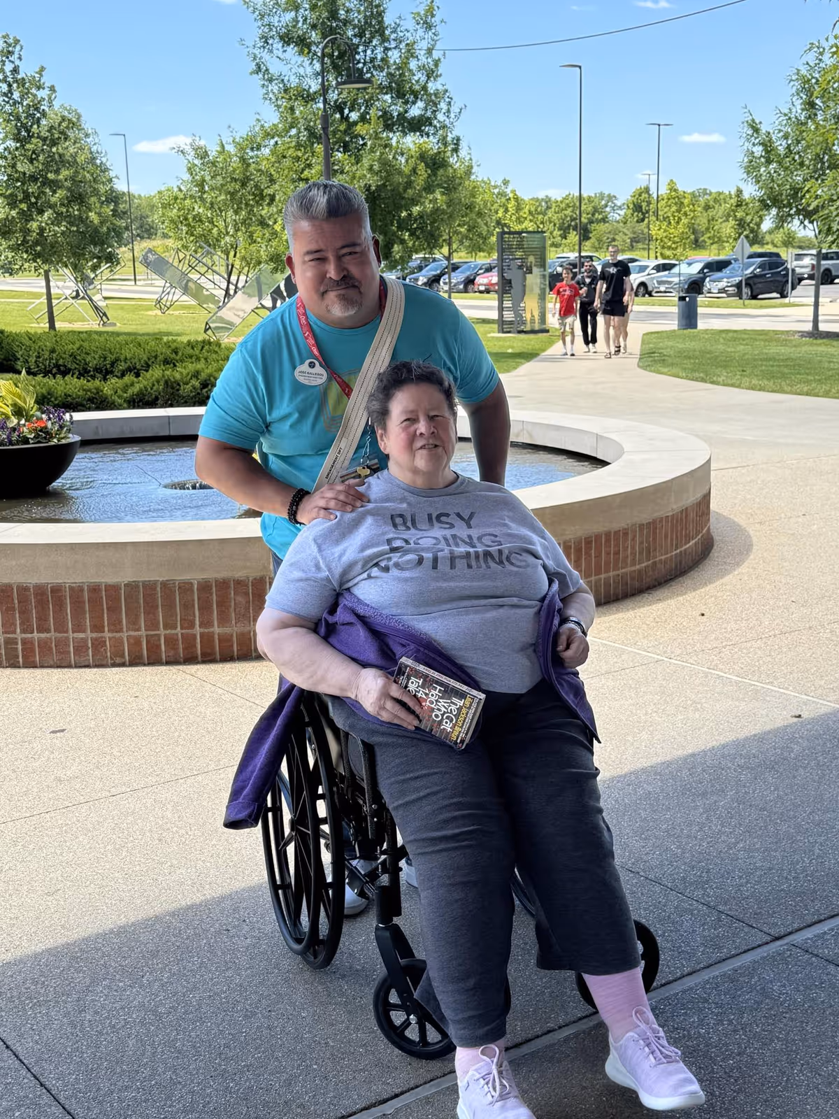 A woman in a wheelchair wearing a gray shirt that says 'BUSY DOING NOTHING' is being pushed by a man in a turquoise shirt. They are outside near a circular water fountain with trees and parked cars in the background. Several people are walking on a pathway behind them.