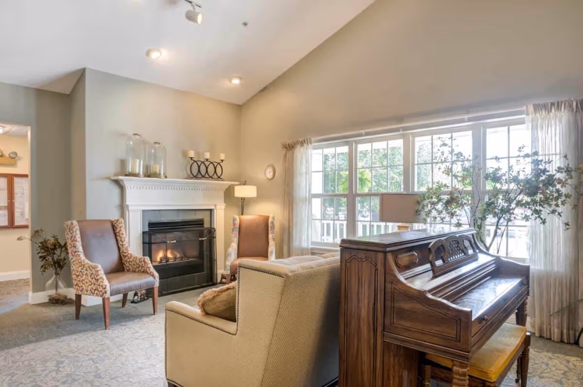 A cozy living room area with a lit fireplace, two upholstered armchairs, a beige sofa, and a wooden piano near a large window with sheer curtains. There are decorative candles on the fireplace mantel and a floor lamp beside one of the chairs.