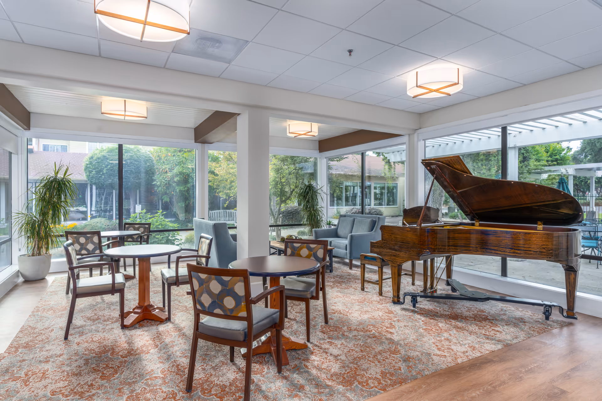 A bright and spacious common area with large windows overlooking a garden. The room features a grand piano, several round tables with patterned chairs, and a seating area with two gray armchairs. The floor is covered with a patterned rug, and there are potted plants near the windows.