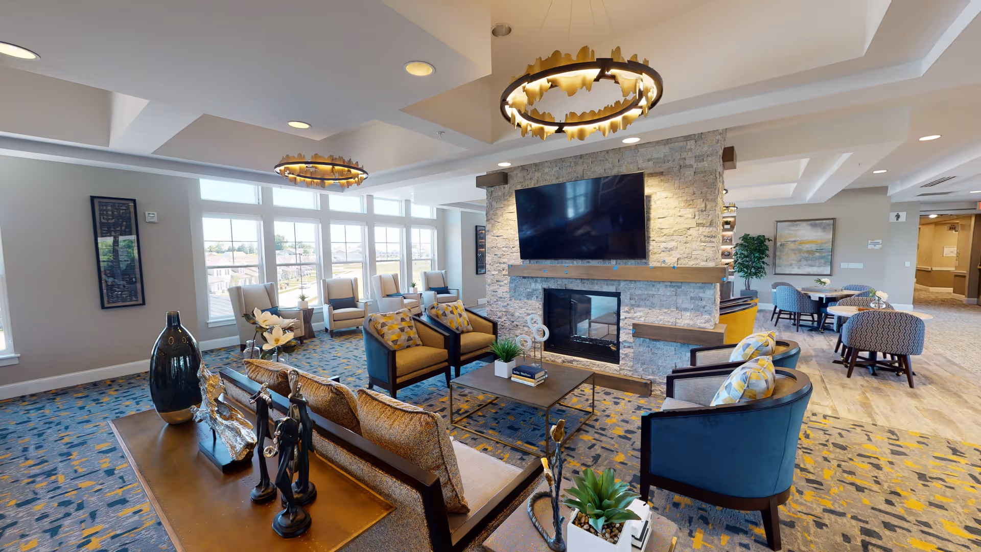 A spacious senior living community lounge with modern decor featuring a stone fireplace with a large flat-screen TV mounted above it. The room has multiple seating options including armchairs, sofas, and small tables with chairs. Large windows allow natural light to fill the space, and decorative light fixtures hang from the ceiling. Various decorative items and plants are placed around the room.