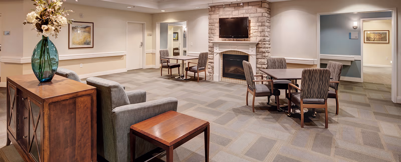 Well-lit lounge area with seating, tables, a stone fireplace with a TV, and a decorative vase on a sideboard.