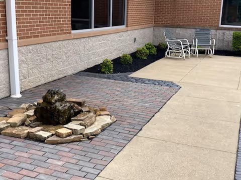 Outdoor patio area with a small rock water feature surrounded by bricks, adjacent to a building with brick and stone walls. There are two metal chairs and a small table placed on the concrete patio near the building wall, with some small green shrubs planted along the edge.