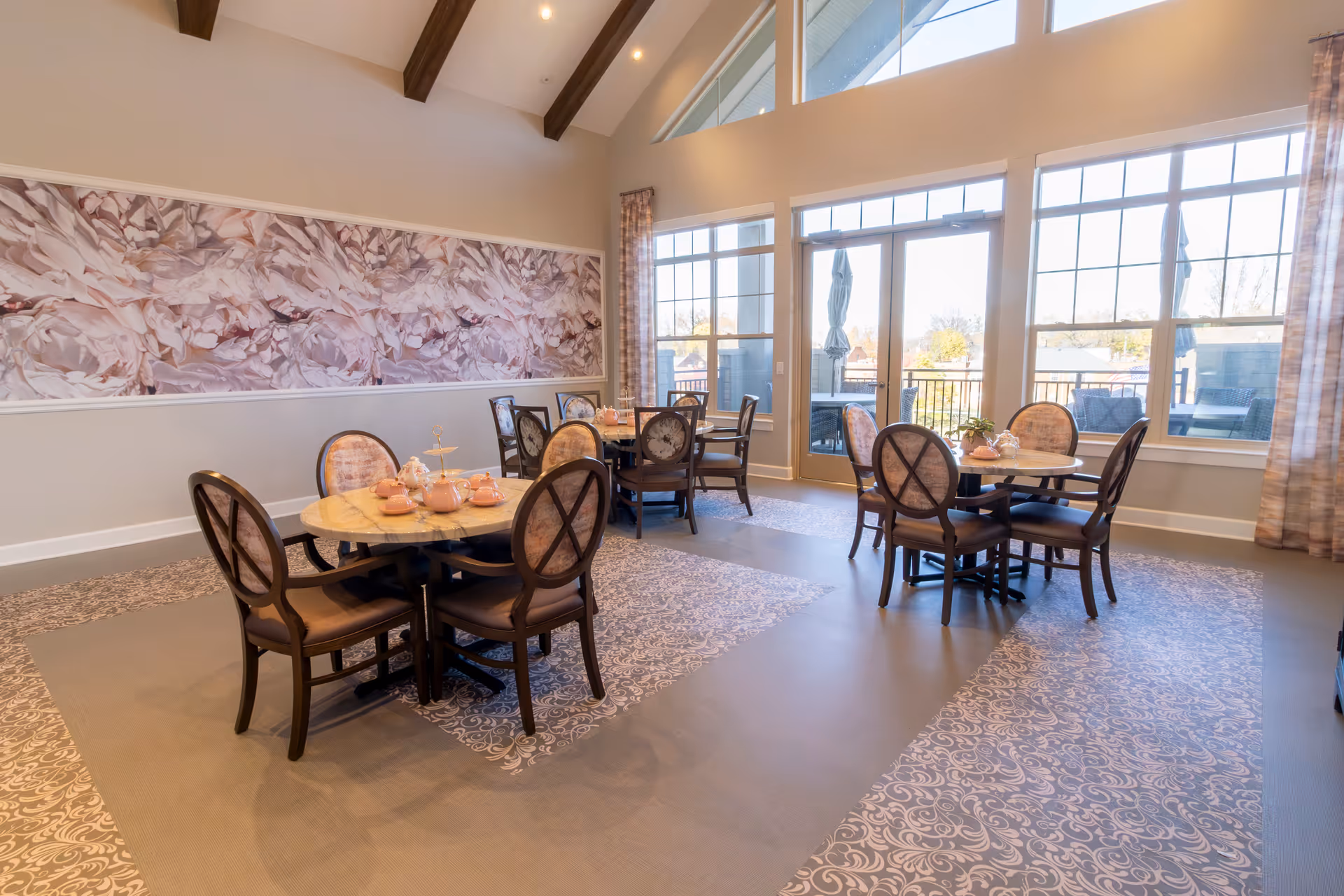 A bright dining room with round tables and cushioned chairs arranged neatly. The room features large windows and a glass door leading outside, allowing natural light to fill the space. The walls are decorated with a large floral mural, and the ceiling has exposed wooden beams. The tables are set with pink teapots and cups.