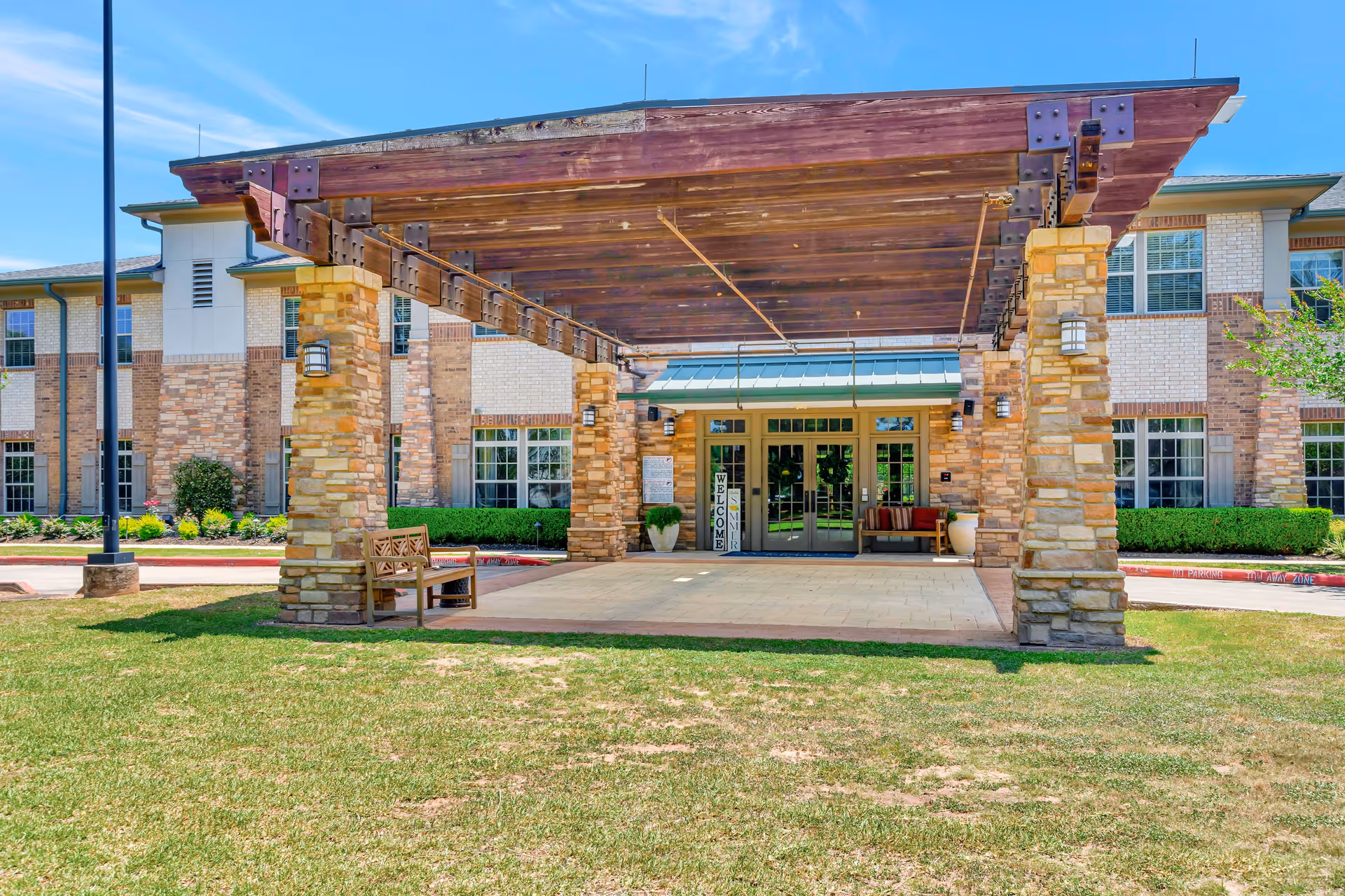 Front entrance of The Avenues of Fort Bend senior living facility featuring a large wooden pergola supported by stone pillars, a bench on the left side, and glass double doors with a welcome sign. The building is two stories with brick and light-colored siding, surrounded by a grassy area and landscaping.