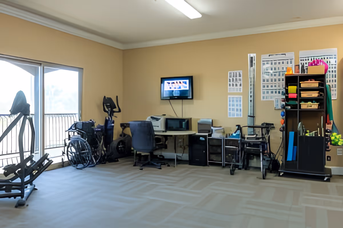 Common area with exercise equipment, several wheelchairs and walkers, a desk with a wall-mounted TV, and storage shelves.