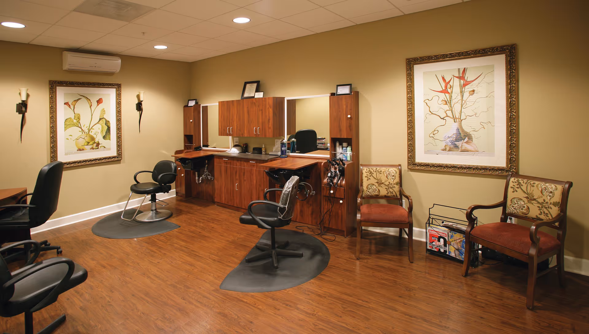 Interior view of a salon area in a senior living facility with wooden flooring, two salon chairs on mats in front of a counter with mirrors and cabinets, two upholstered chairs with floral patterns, framed botanical artwork on the walls, and warm lighting.