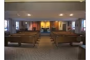 Interior view of a chapel or worship room with rows of wooden pews facing an altar at the front. The room has carpeted flooring, soft lighting from ceiling fixtures, and windows along the side walls.