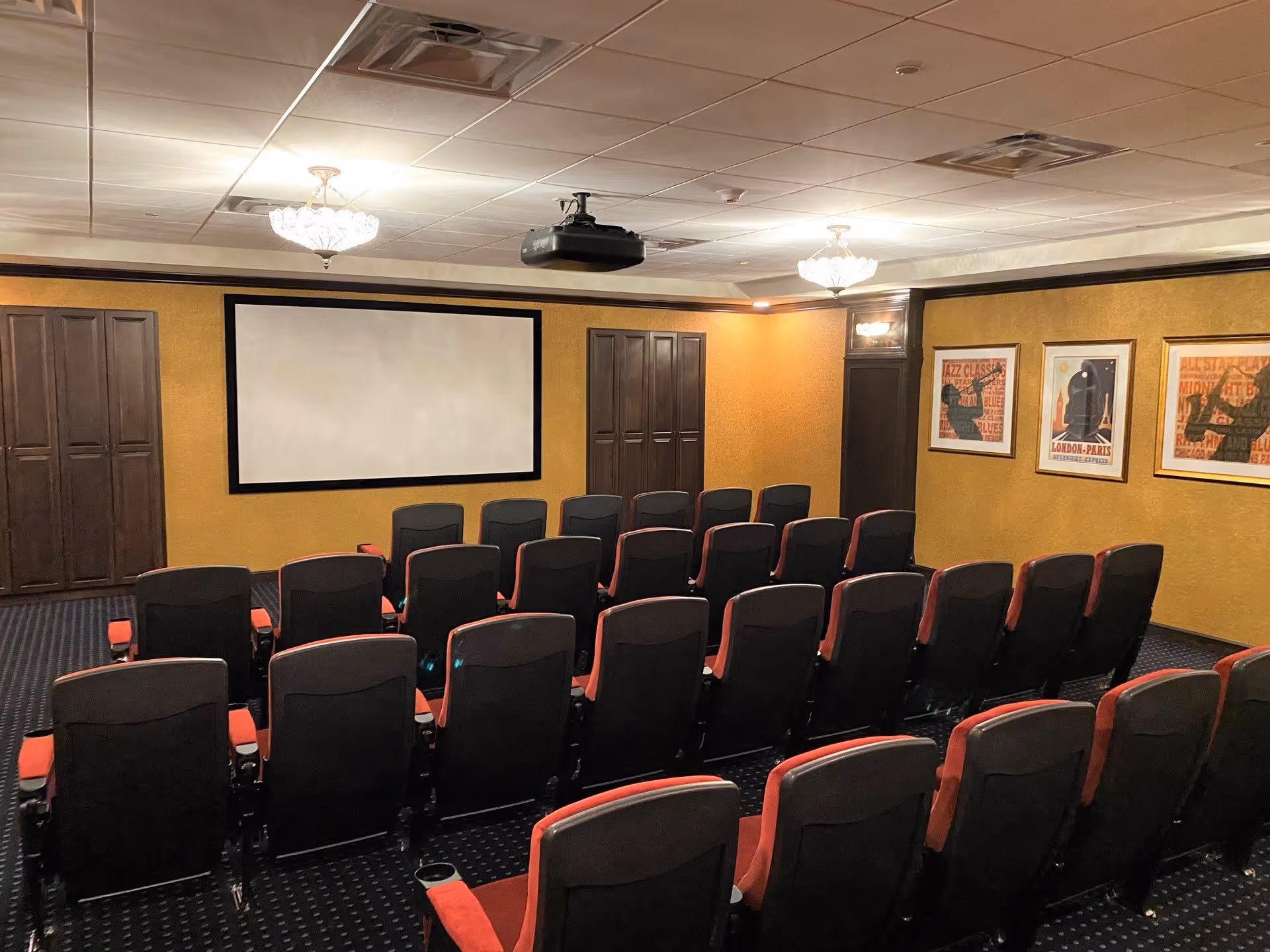 A small theater room with three rows of red and black cushioned seats facing a large blank projection screen on a yellow wall. The room has a dark carpet with a subtle pattern, two chandeliers on the ceiling, and framed artwork on the right wall. There are wooden cabinets on either side of the screen.