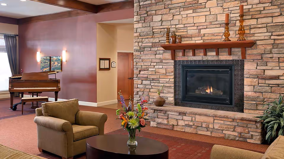 A cozy living room area featuring a stone fireplace with a fire burning, a wooden mantel decorated with candles and small vases, a round coffee table with a colorful flower arrangement, an armchair, and a grand piano in the background near a window with curtains.