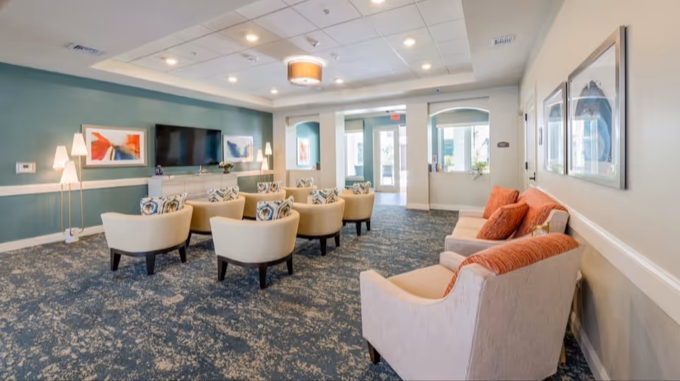 A spacious and well-lit living room area in a senior living facility with multiple beige armchairs arranged in rows facing a wall-mounted flat-screen TV. The room has a blue patterned carpet, teal and white walls, modern floor lamps, and framed abstract artwork. There is a beige sofa with orange cushions along one wall and large windows near the entrance letting in natural light.