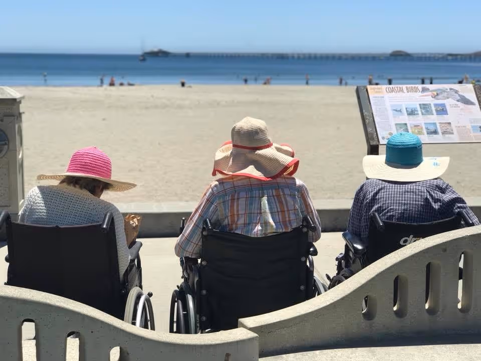 Three elderly individuals in wheelchairs wearing sun hats sitting on a concrete bench facing a sandy beach and ocean with a pier in the distance.