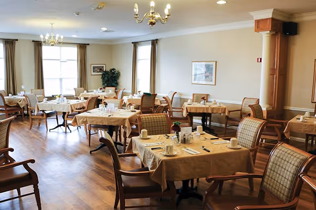 A bright dining room with multiple tables covered in beige tablecloths, each set with white cups, plates, and silverware. The room has wooden flooring, cushioned chairs with wooden frames, large windows with curtains, chandeliers hanging from the ceiling, and framed artwork on the walls.
