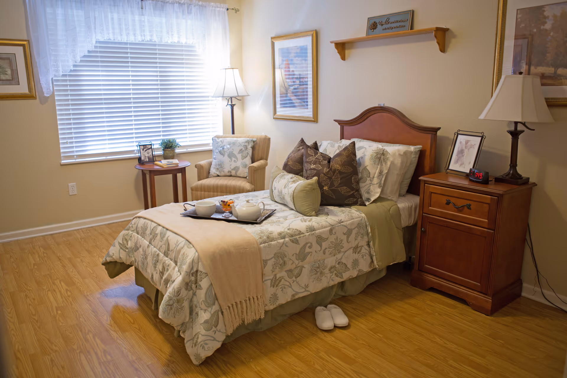 A cozy bedroom with a single bed covered in floral bedding and multiple pillows. A tray with a teapot, cup, and small bowl is placed on the bed. Next to the bed is a wooden nightstand with a lamp, framed picture, and digital clock. A comfortable armchair with a cushion and a small round table with a plant and photo frame are positioned near a window with white blinds and sheer curtains. The room has wooden flooring and soft, warm lighting.