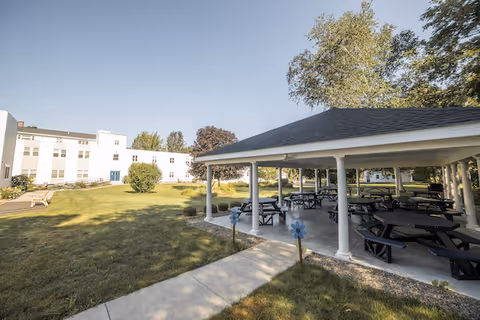 Covered outdoor pavilion with picnic tables on a lawn next to a multi-story senior living building under a clear sky.