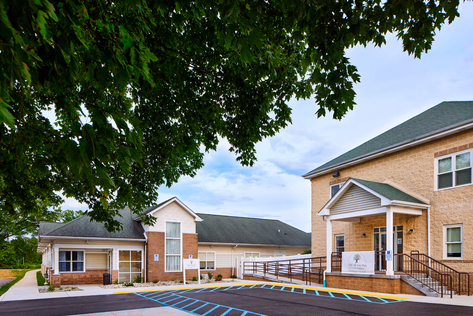 Exterior view of The Seasons of Saxonburg senior living facility showing two connected buildings with beige and light brown walls, green roofs, a wheelchair ramp, and a parking area with handicap spaces. Large green tree branches frame the top left of the image under a partly cloudy sky.