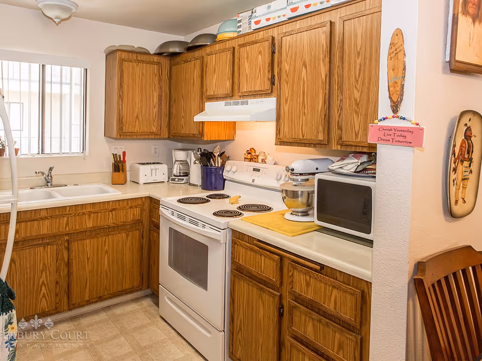 A kitchen with wooden cabinets, a white electric stove with four burners, a white microwave, a stand mixer, a coffee maker, a toaster, and various kitchen utensils. There is a window with bars above the sink, and a small decorative sign on the wall that reads 'Cherish Yesterday Live Today Dream Tomorrow.'
