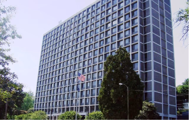Large multi-story rectangular building with a grid of windows, flagpoles and trees in front.
