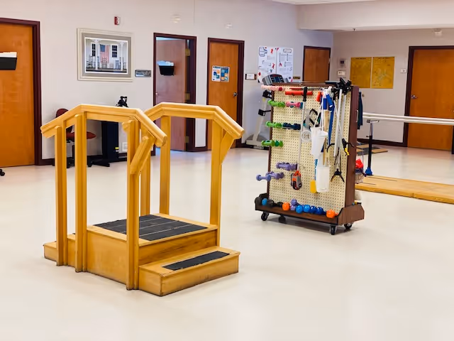 Interior room with a wooden physical therapy step platform and a mobile rack holding various exercise equipment such as dumbbells, resistance bands, and balls. The room has several closed wooden doors, bulletin boards, and a framed picture on the wall.