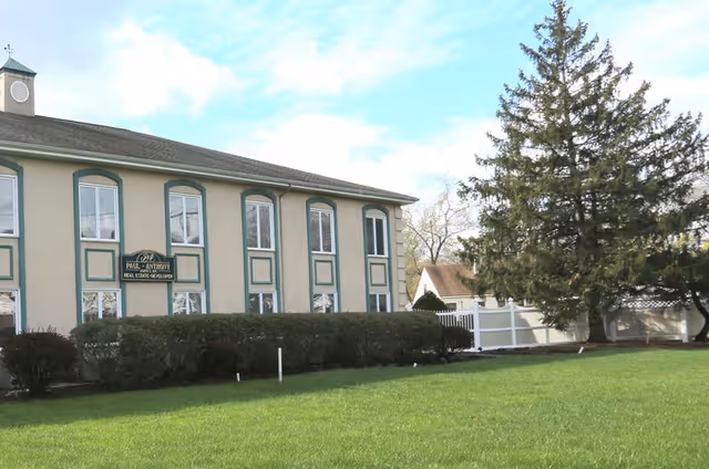 Front exterior of a two-story beige building with green trim, manicured lawn, shrubs, and a large evergreen tree.