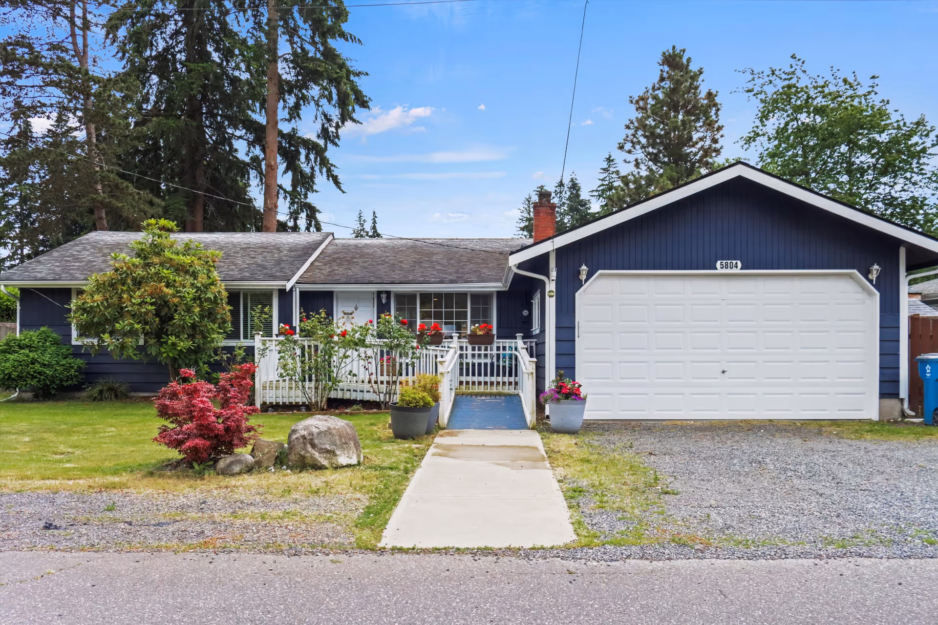 Front exterior view of a single-story blue house with a white garage door, a concrete walkway leading to a small porch with white railings, and various plants and flowers in the front yard under a partly cloudy sky.