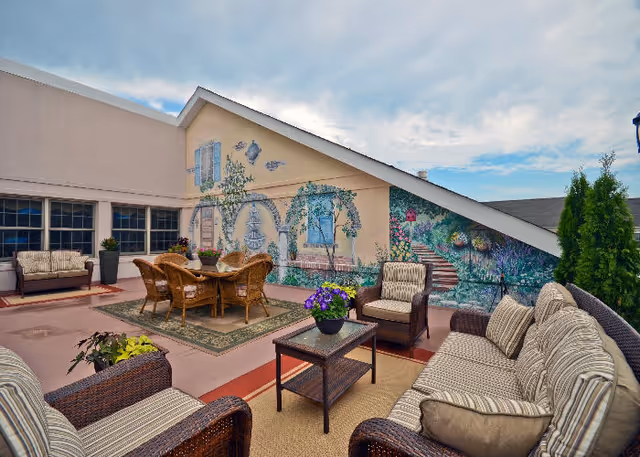 Outdoor patio area with wicker furniture including sofas, armchairs, and a table with chairs. The area features a mural painted on a slanted wall depicting a garden scene with arches, plants, and flowers. Potted plants and a clear sky with some clouds are visible.