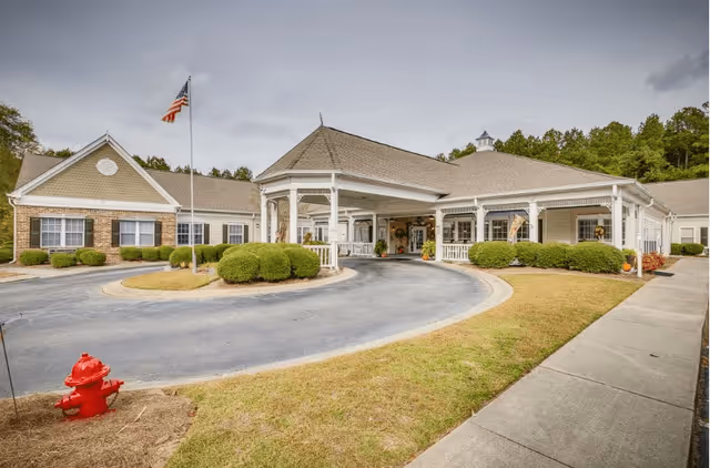Front exterior view of The Legacy of Orangeburg facility showing a single-story building with a covered entrance, manicured bushes, an American flag on a flagpole, a red fire hydrant, and a curved driveway.