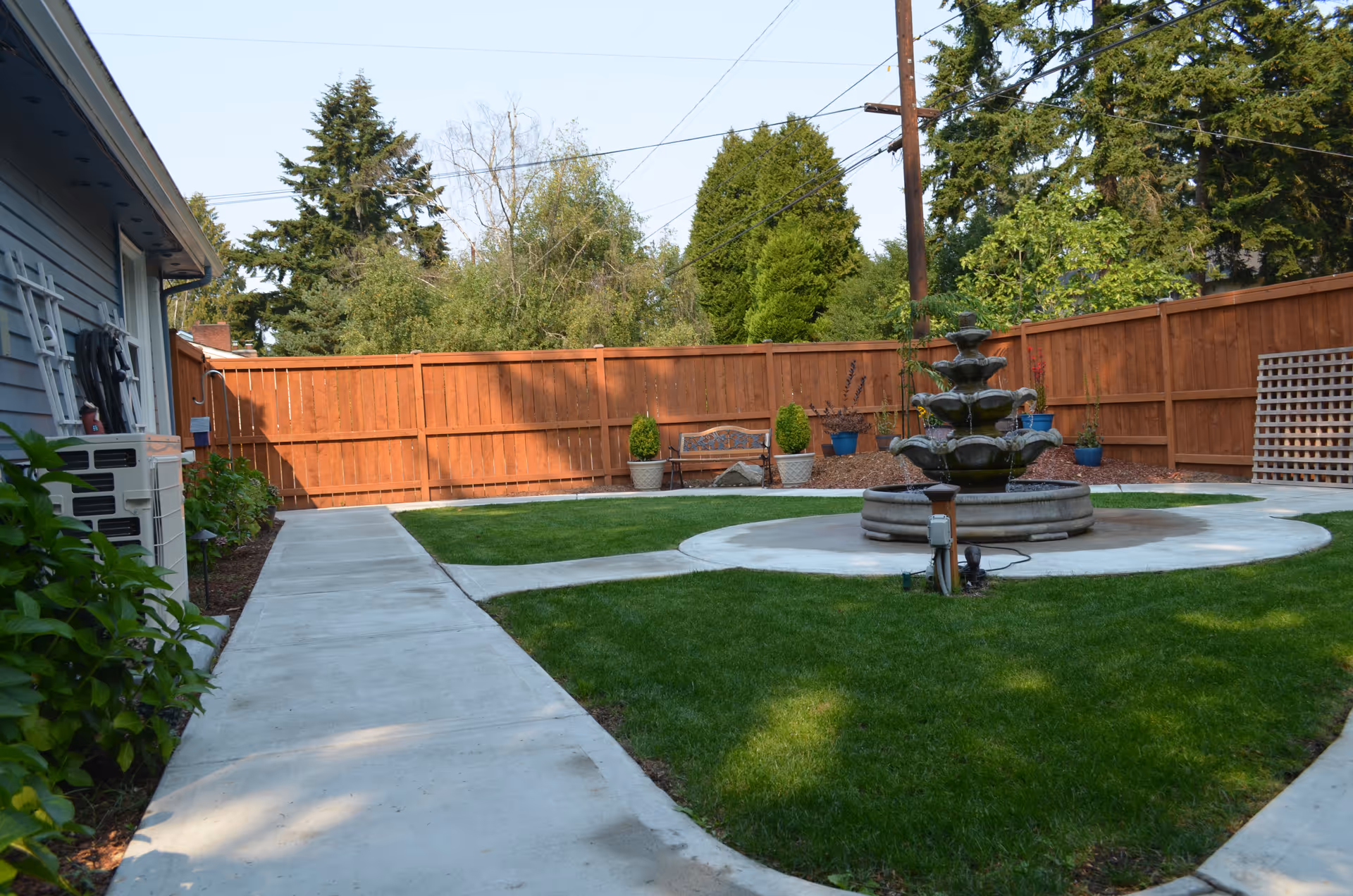 Backyard courtyard with a central multi-tiered fountain surrounded by a circular concrete path, green lawn, wooden fence and a side walkway by the house.