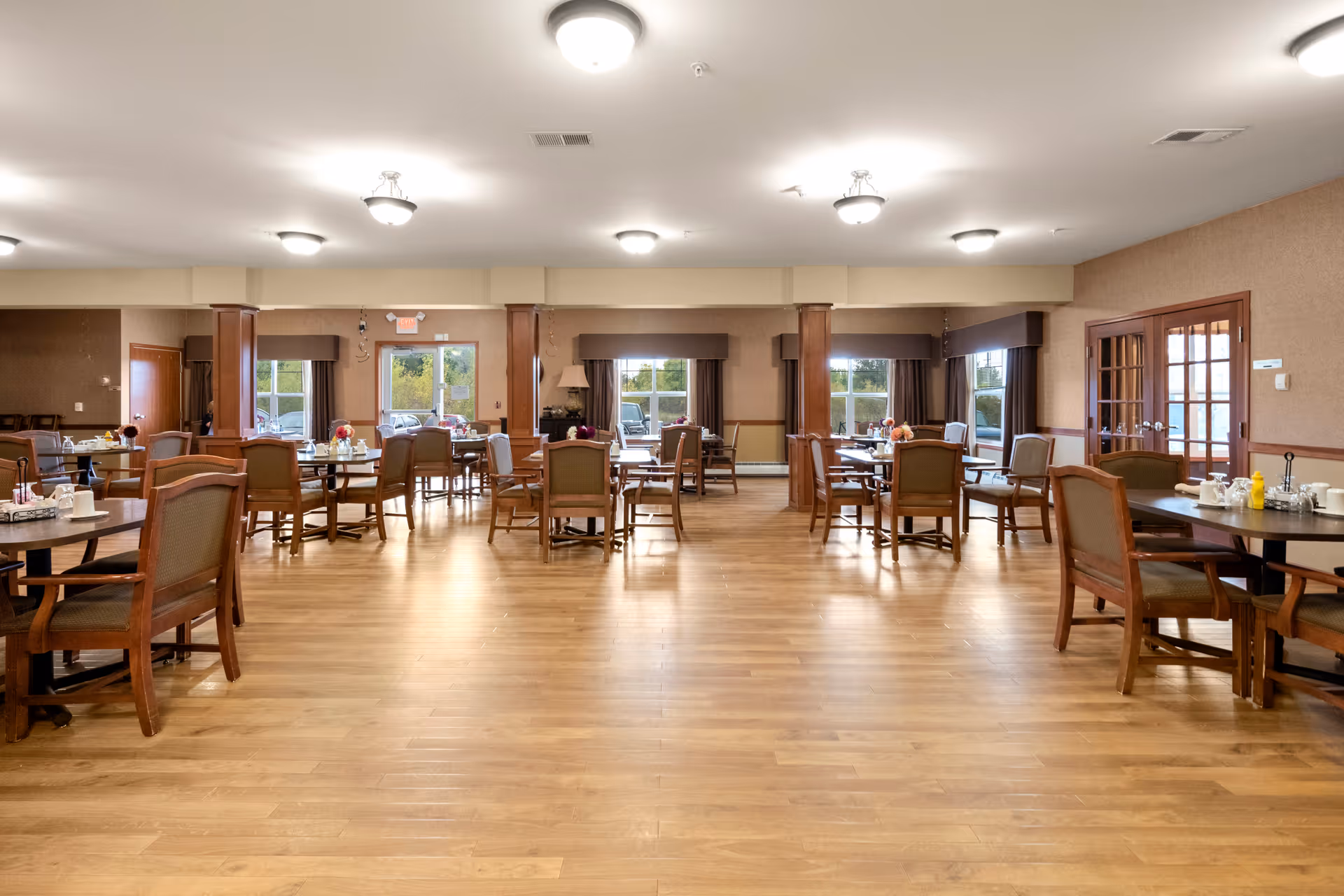A spacious dining room in a senior living facility with multiple round wooden tables and cushioned chairs arranged neatly. The room has large windows with brown valances allowing natural light to brighten the space. The floor is wooden, and the ceiling has several light fixtures evenly spaced. There are doors and columns visible along the walls.